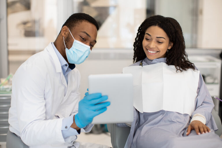 Dentist in a face mask and blue gloves showing a tablet to a smiling patient in a dental chair, illustrating HotDoc vs Healthengine online booking